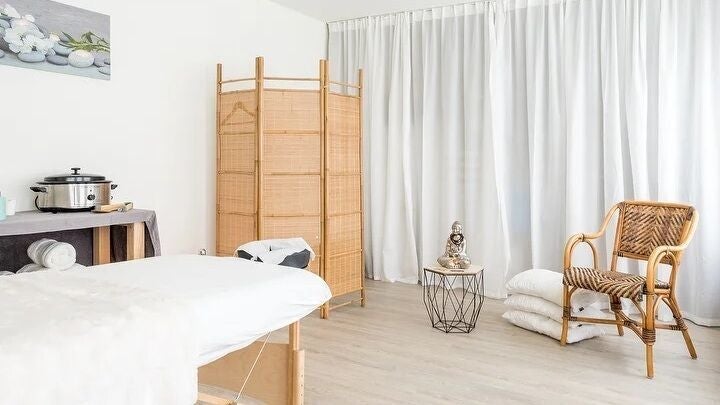 De lichte behandelruimte met massagetafel, rotan kamerscherm, rotan stoel en witte gordijnen